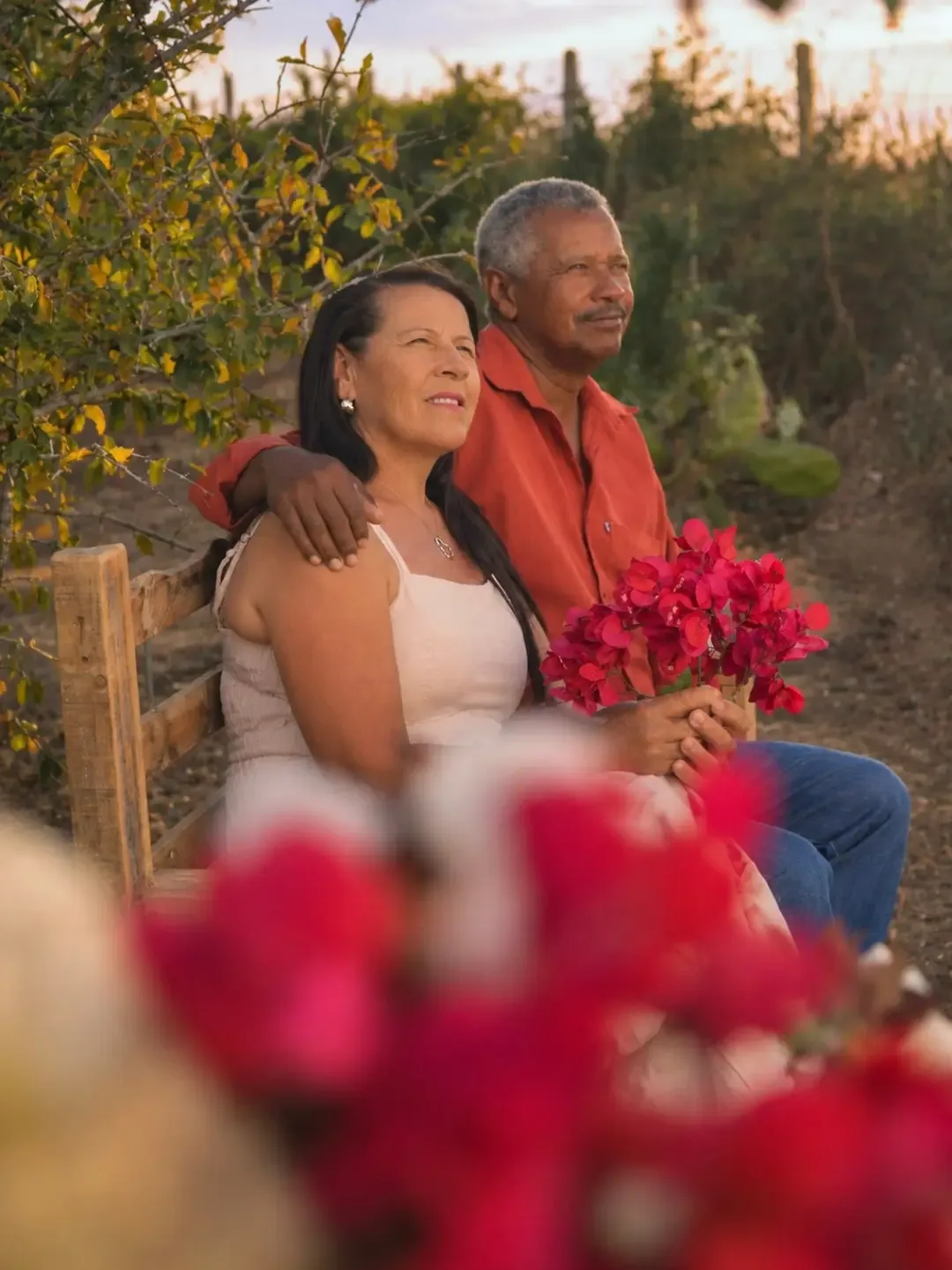 Casal sentado ao ar livre, com flores cor-de-rosa em primeiro plano.